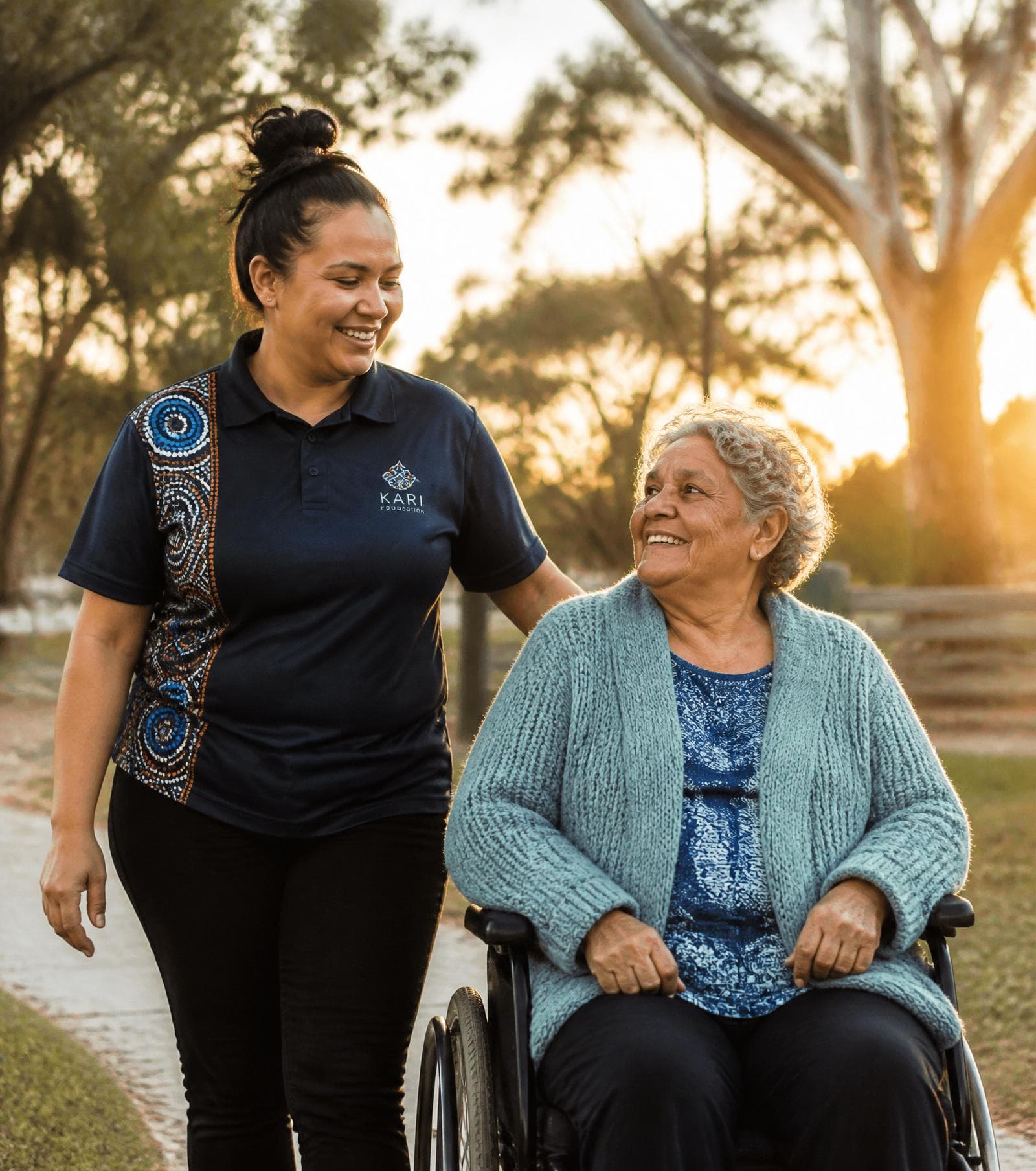 A support worker walks alongside an older Aboriginal woman in a wheelchair through a sunlit park